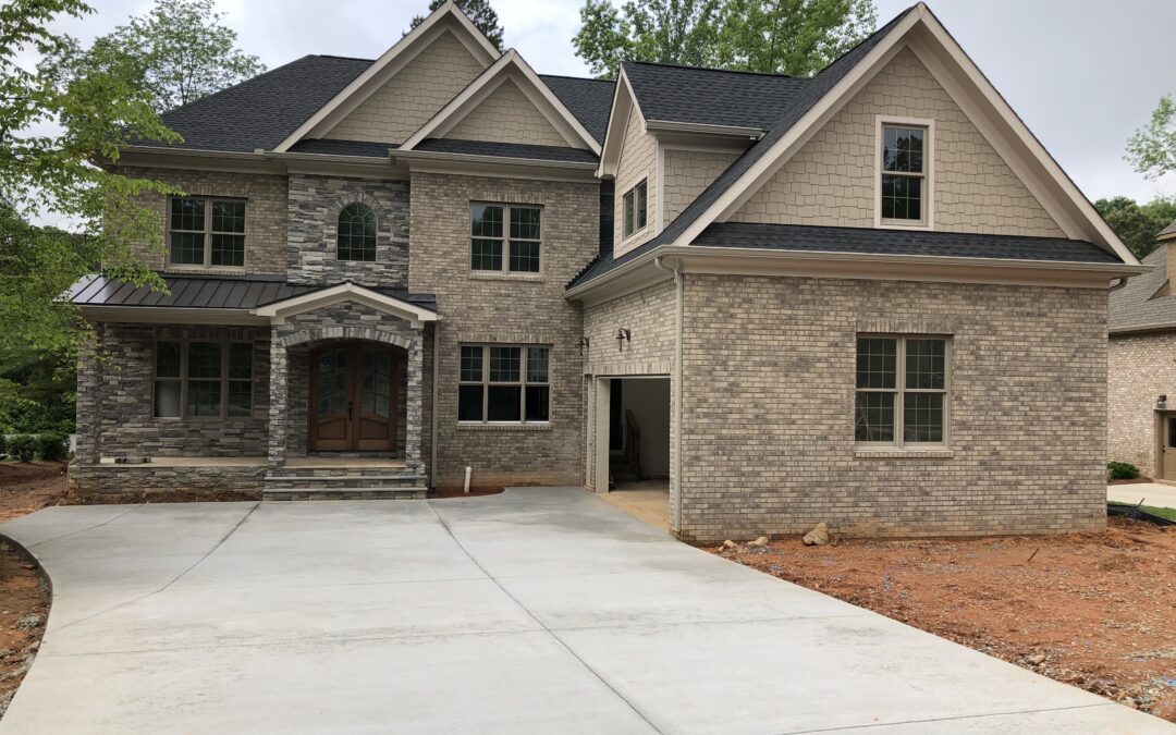 Two-story house with a mix of brick and stone facade, double-front doors, and an attached garage on a freshly paved driveway. Surrounding trees in the background.