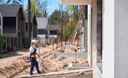 Construction worker in a helmet and vest walks along a path at a residential construction site, with partially built houses and scaffolding in view.