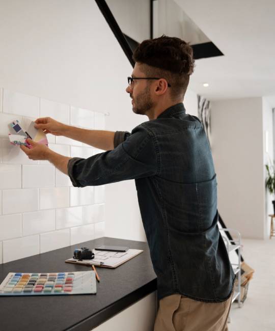 A man in glasses is holding color swatches against a white tiled wall in a modern room. A color palette and clipboard are on the counter beside him.