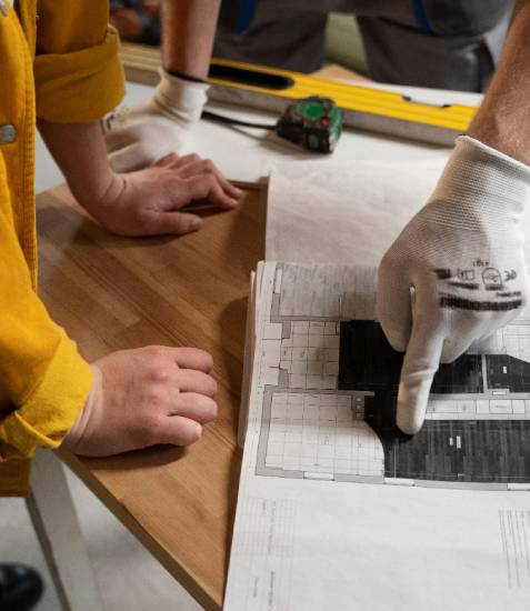 Two people examining architectural blueprints on a wooden table, with a tape measure and level nearby.