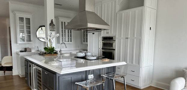 Modern kitchen with a center island featuring a stove and range hood, glass-front cabinets, a round mirror, and two transparent bar stools. White and gray color scheme.