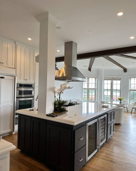 Modern kitchen with a large island, stainless steel appliances, white cabinets, and a chimney hood. A potted orchid sits on the island, and natural light streams through large windows.