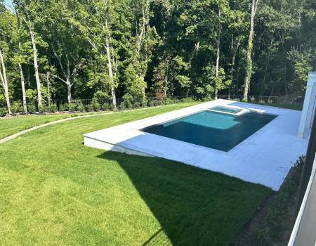 A rectangular pool with a small attached spa is situated in a well-manicured grassy yard surrounded by trees.