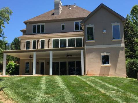 A large, two-story house with a sloped roof, multiple windows, and a covered patio. The lawn is freshly mowed, and trees are visible in the background.