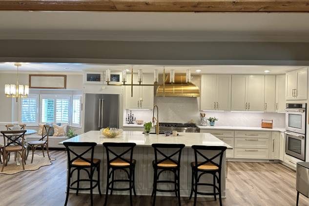 Modern kitchen with a large island, barstools, stainless steel appliances, and a chandelier. Dining area with a round table and chairs is visible in the background.