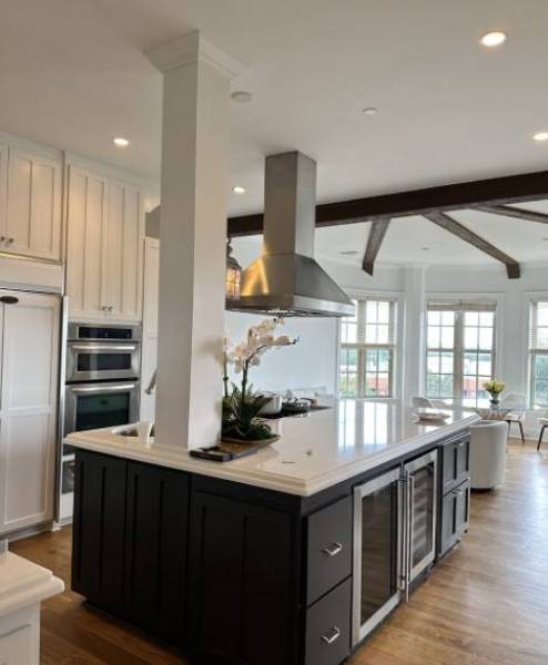 A modern kitchen with a large island, stainless steel appliances, pendant lighting, and wooden beams on the ceiling. A vase with white flowers is on the island.