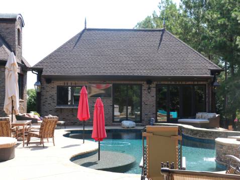 A luxurious backyard view with a brick house, red umbrellas, outdoor furniture, and a pool surrounded by trees.