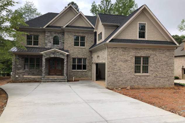 Two-story brick house with a gabled roof, stone accents, and a large driveway. The front features a wooden double door entrance and an open garage. Trees are visible in the background.