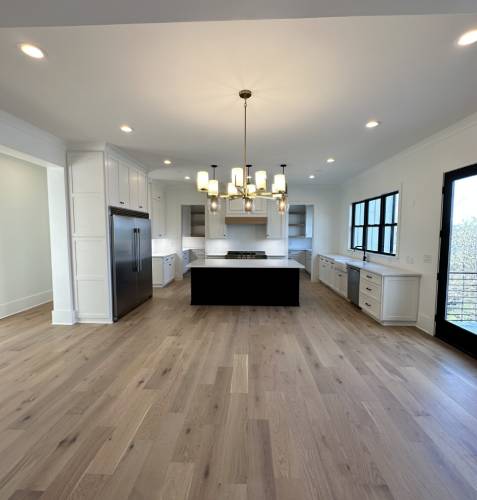 Spacious modern kitchen with hardwood floors, central island, stainless steel fridge, white cabinets, and a chandelier. Large windows and a glass door provide natural light.