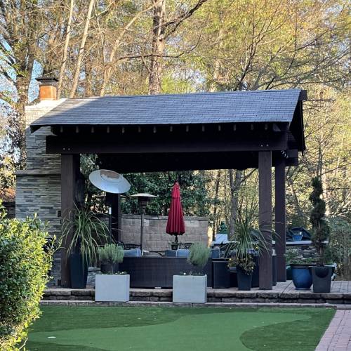 A wooden pergola with a grill and chimney stands in a garden. There are potted plants and a red umbrella beside it, surrounded by trees and greenery.