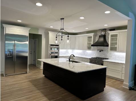 A modern kitchen with a large island, white cabinets, black range hood, stainless steel appliances, and wood flooring. Pendant lights hang above the island.