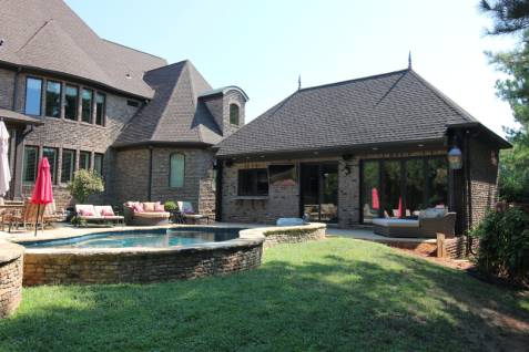 A brick house with a dark roof, a small swimming pool, and patio furniture in the backyard.