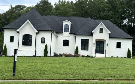 A white modern house with a dark roof and multiple gabled sections, surrounded by a neatly trimmed lawn and small, newly planted shrubs.