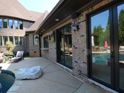 A brick house with large windows overlooks a patio featuring a pool, lounge chairs, and outdoor seating.