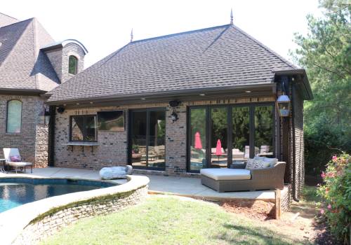 A brick house with a pointed roof features large windows, a cushioned outdoor sofa, and a pool nearby, surrounded by a grassy area and trees.