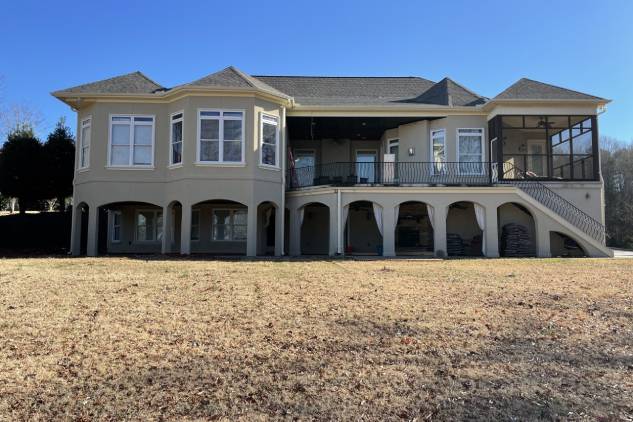 A two-story house with large bay windows and an outdoor staircase leading to a covered patio. The lawn in the foreground is brown and dry.