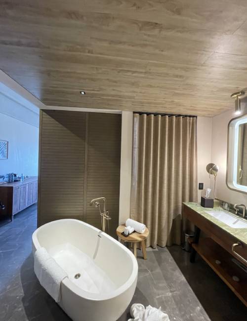 Modern bathroom with a freestanding oval bathtub, a wooden stool with towels, a large mirror, and a sink on a wooden counter. Curtains cover a section of the wall.