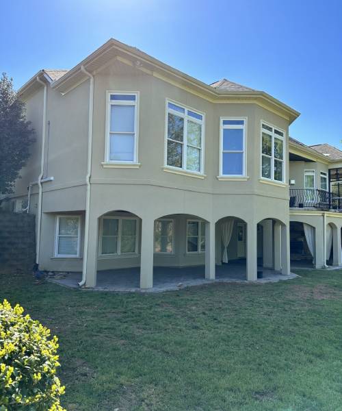 Two-story beige house with arched covered patio and large windows, surrounded by grass and bushes, under a clear blue sky.