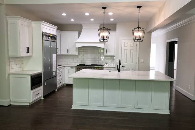 Modern kitchen with white cabinets, marble countertops, and stainless steel appliances. Two pendant lights hang above the island with a dark wood floor.