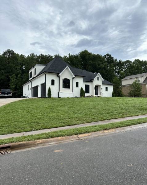 A white two-story house with a sloped black roof sits on a grassy hill. There's a driveway on the left and trees in the background under a cloudy sky.